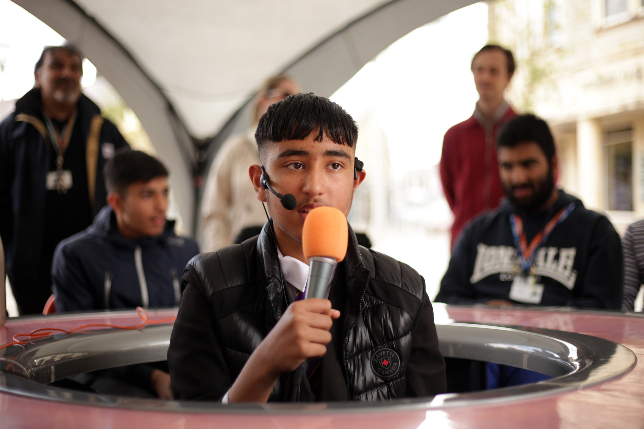 A young man holds a microphone and sits in the middle of a round table, looking outwards and those sitting around the table. 