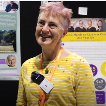 Dr Liz Williamson a woman with short pink hair smiles off camera standing in front of posters at Conference