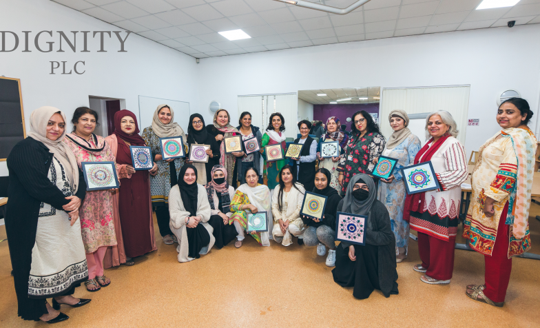 A group of women stand in a semi circle holding paper based art work. In the top left hand corner of the image, is a grey logo that reads 'Dignity PLC'
