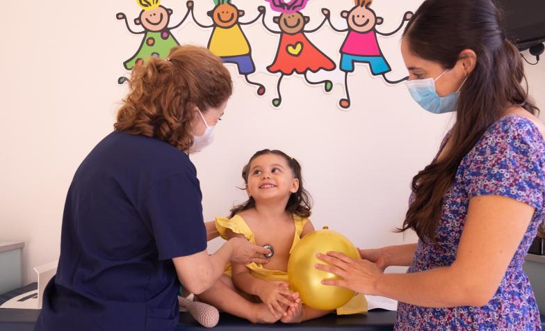 Clinician in navy scrubs uses stethoscope on a smiling child in a yellow dress. 