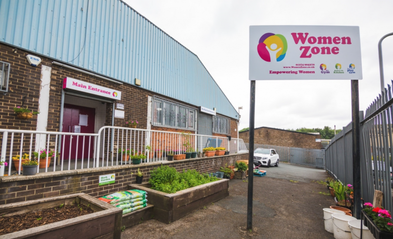 the outside of a brick building, which has a sign that reads 'WomenZone' on it. Infront of the building, their are raised flower beds. The sky is grey.