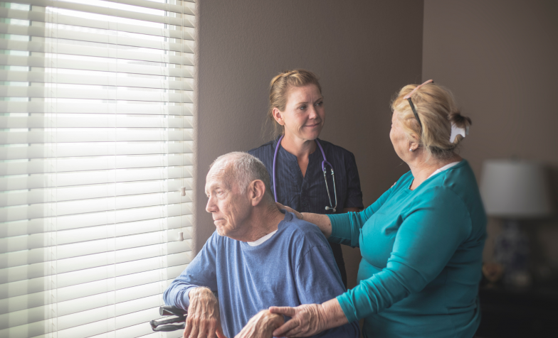 A man in a wheelchair looks out the window with carer and a nurse behind