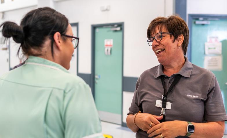 Two women talking to each other in a clinical setting. On the left, one woman is wearing pale green scrubs. On the right, a woman is wearing a grey t-shirt which says 'volunteer'.