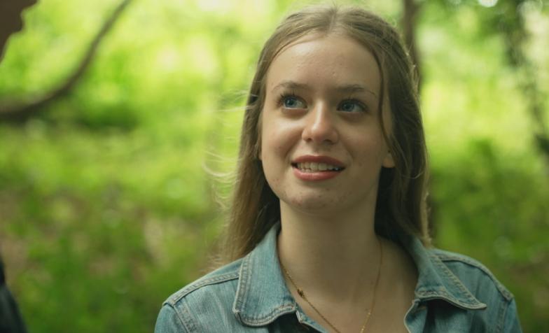 A teenager with long, brunette hair looks up smiling. She is outdoors among the trees of a forest.