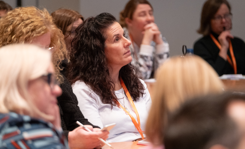 A woman with long dark hair sitting in a crowded room at a conference event.