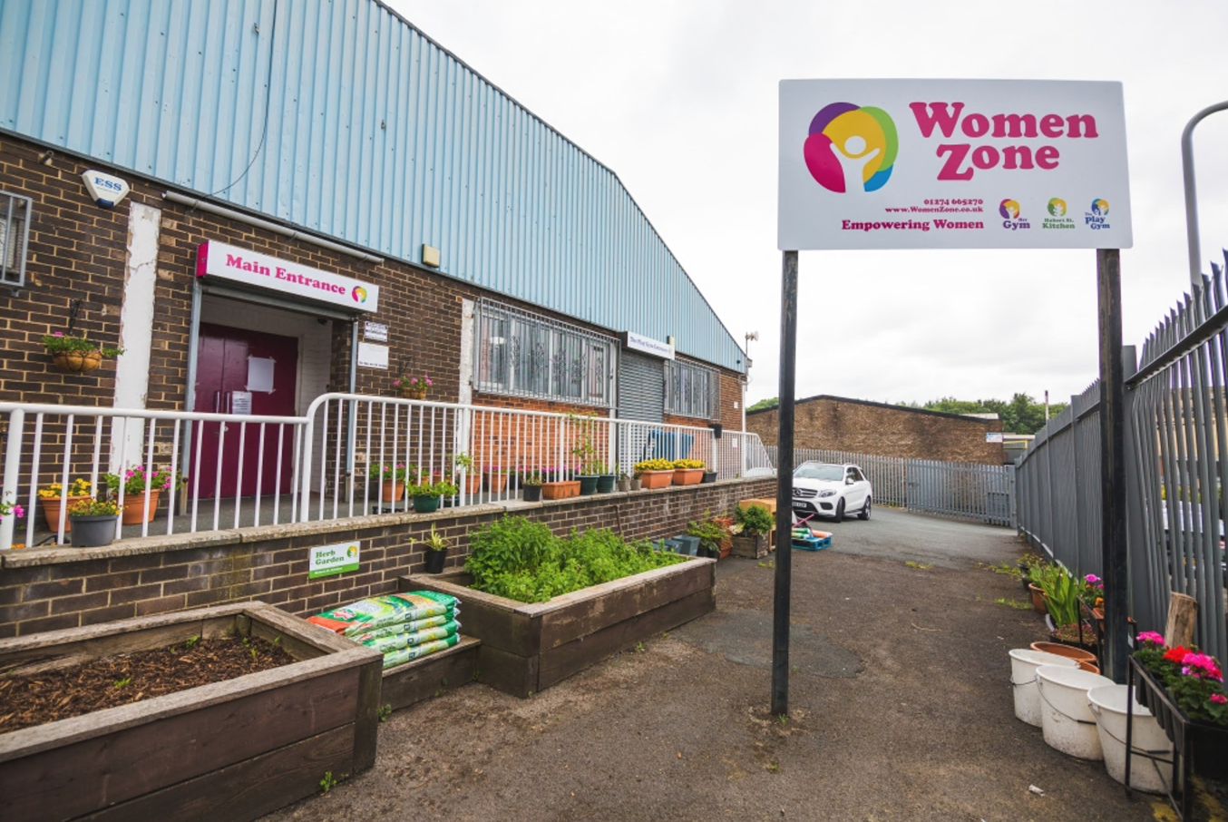 the outside of a brick building, which has a sign that reads 'WomenZone' on it. Infront of the building, their are raised flower beds. The sky is grey.