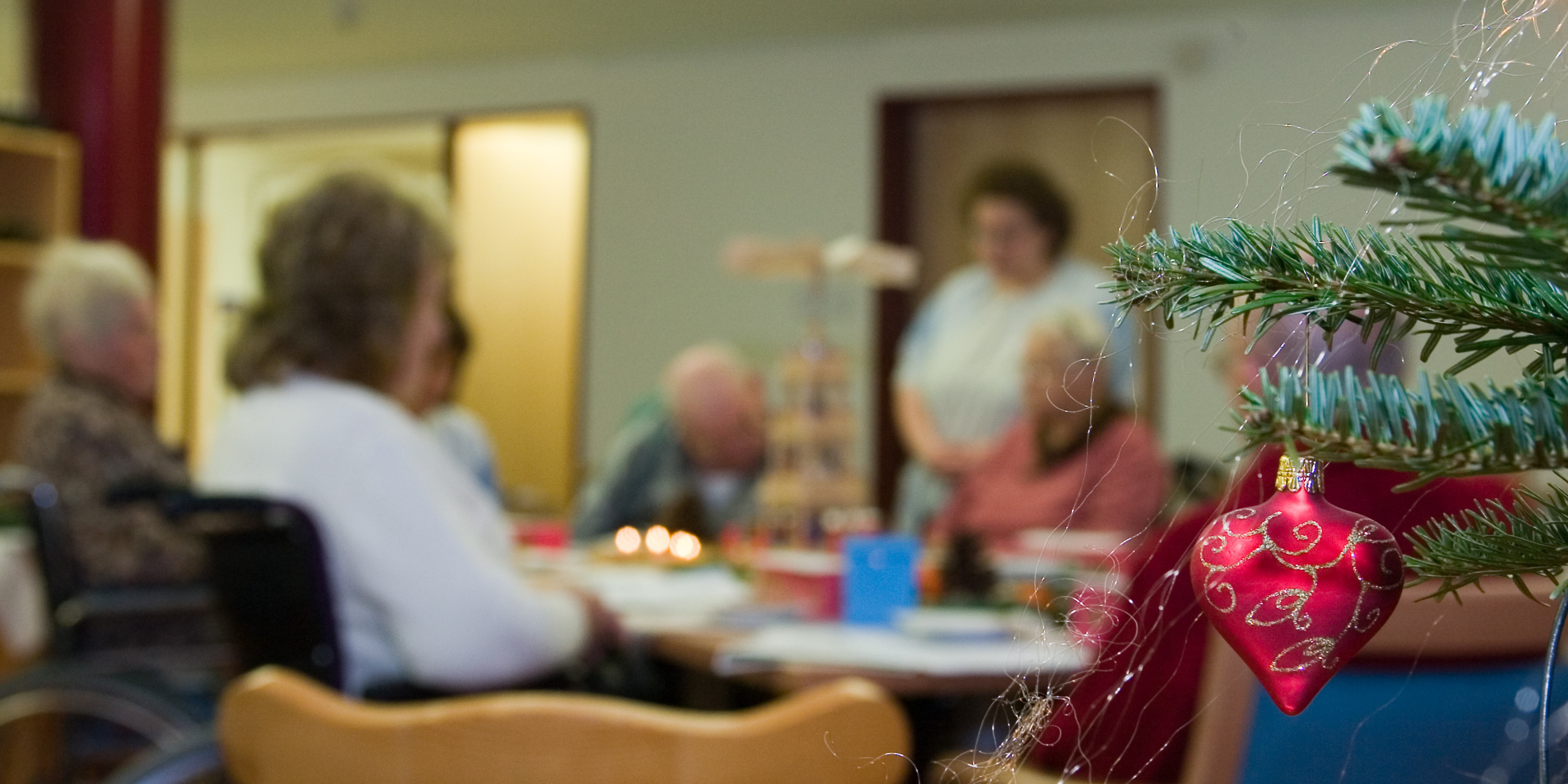 A group of elderly people sitting around a table, participating in an activity, in the foreground there is a Christmas tree with a red bauble on it