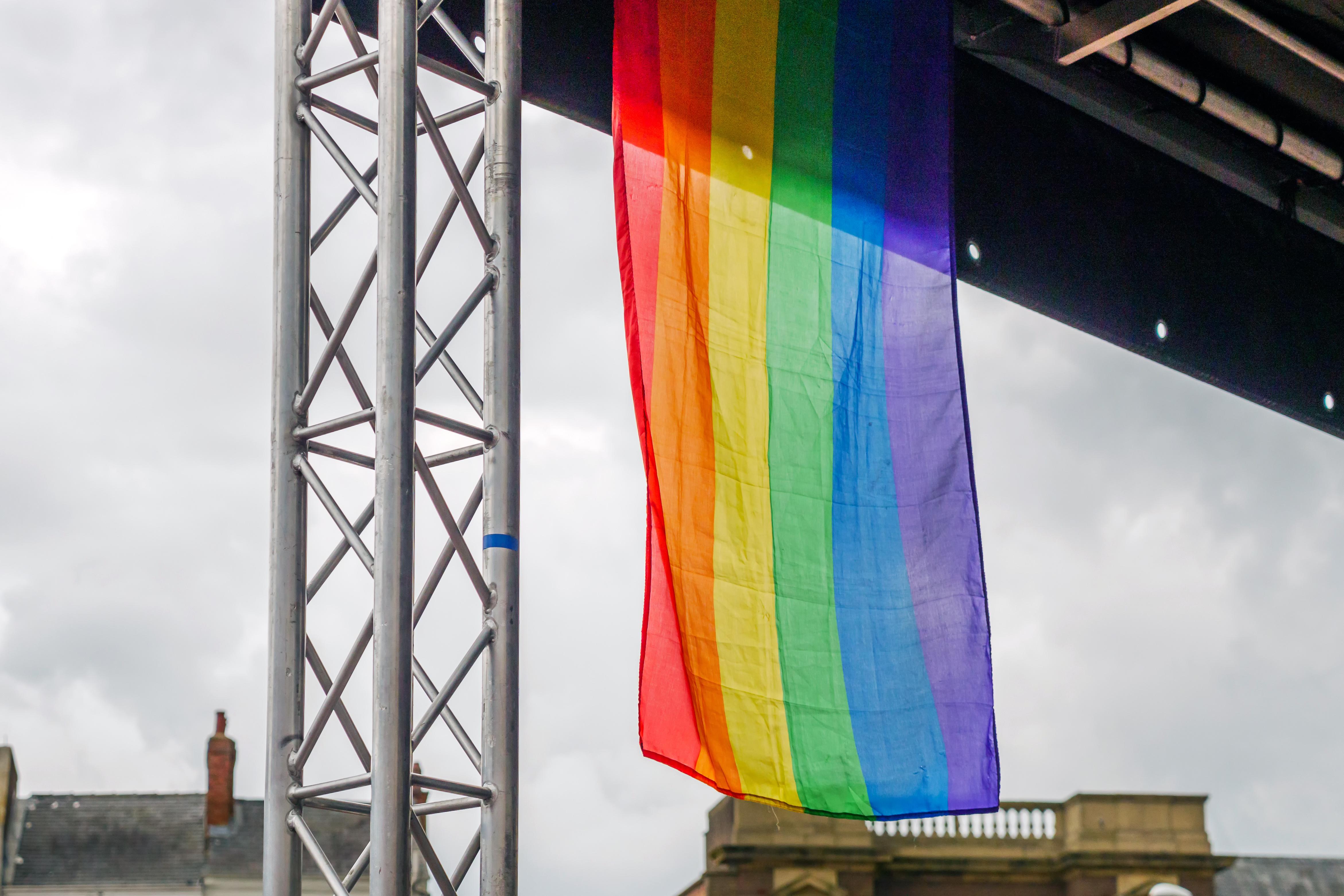 An LGBTQ+ flag hangs from the structure of a stage, outdoors