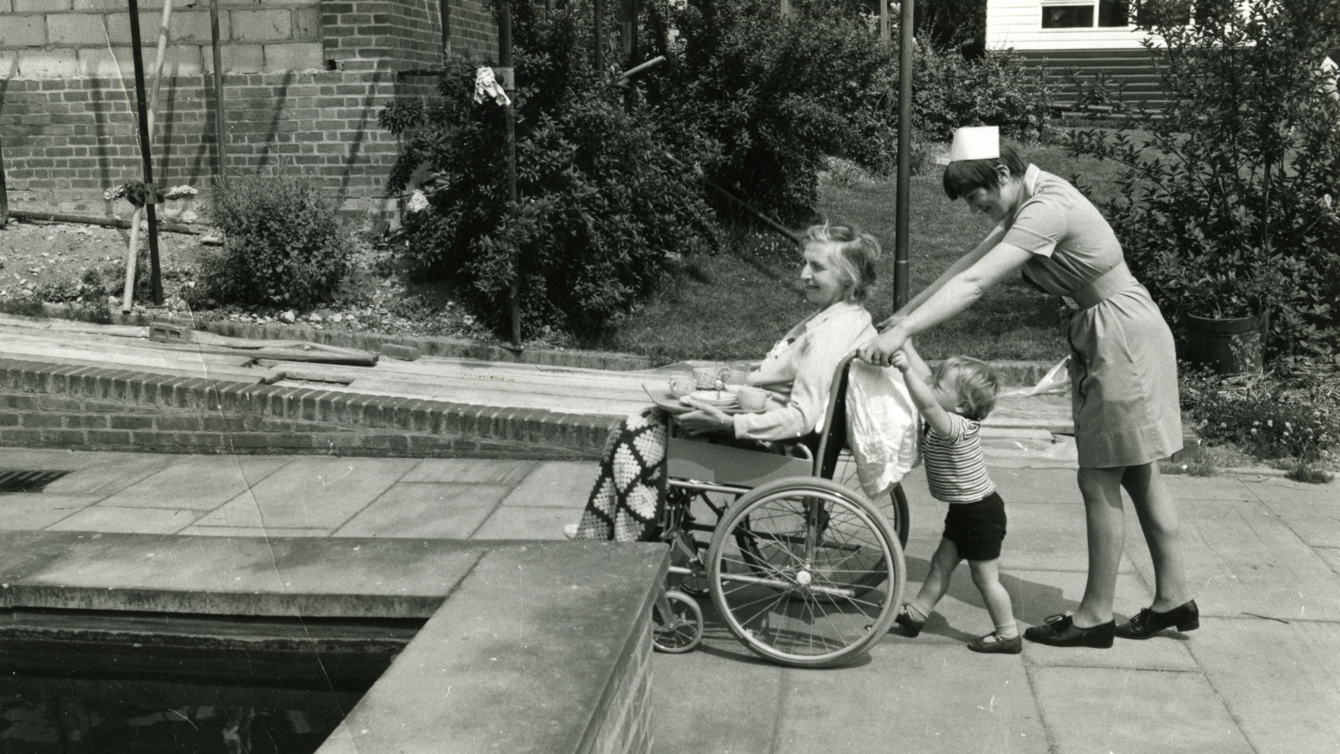 Patient in a wheelchair being pushed by a nurse. A child is attempting to help push the wheelchair.