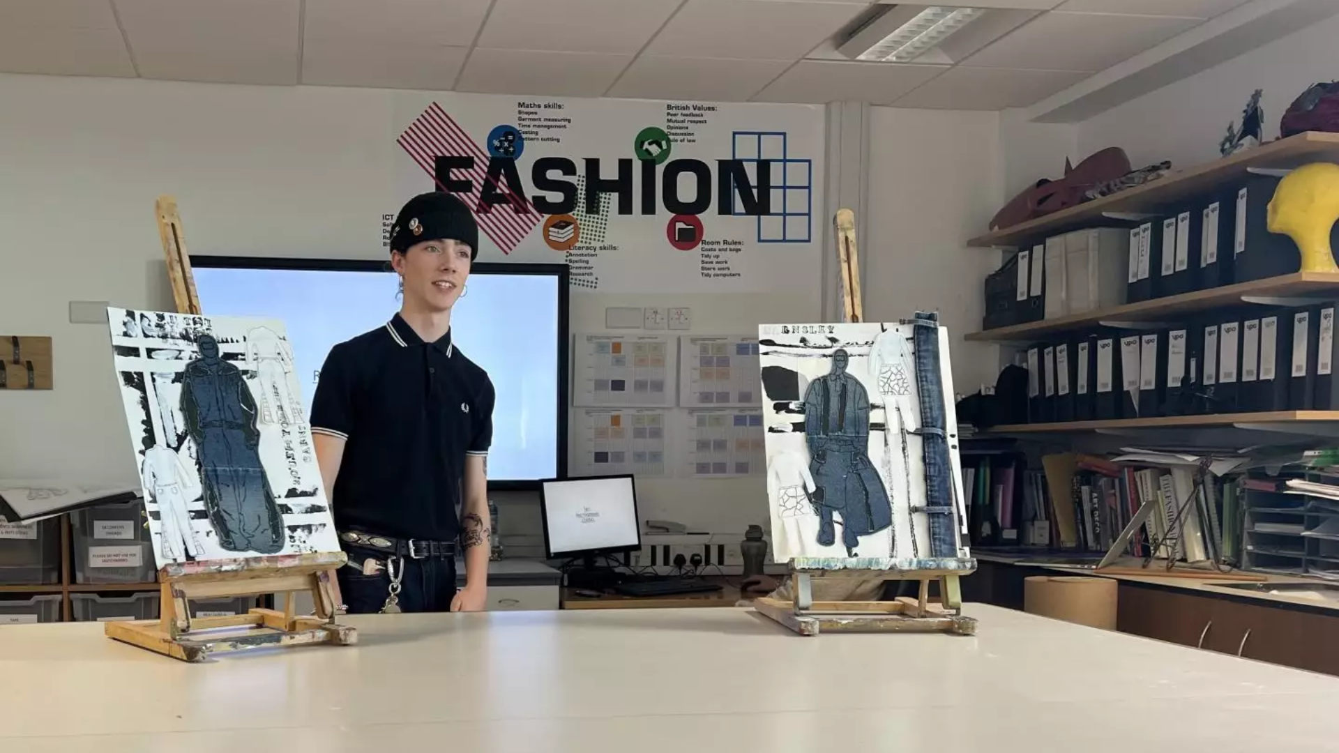 A teenage boy standing at the front of the classroom, presenting canvases with outfit designs