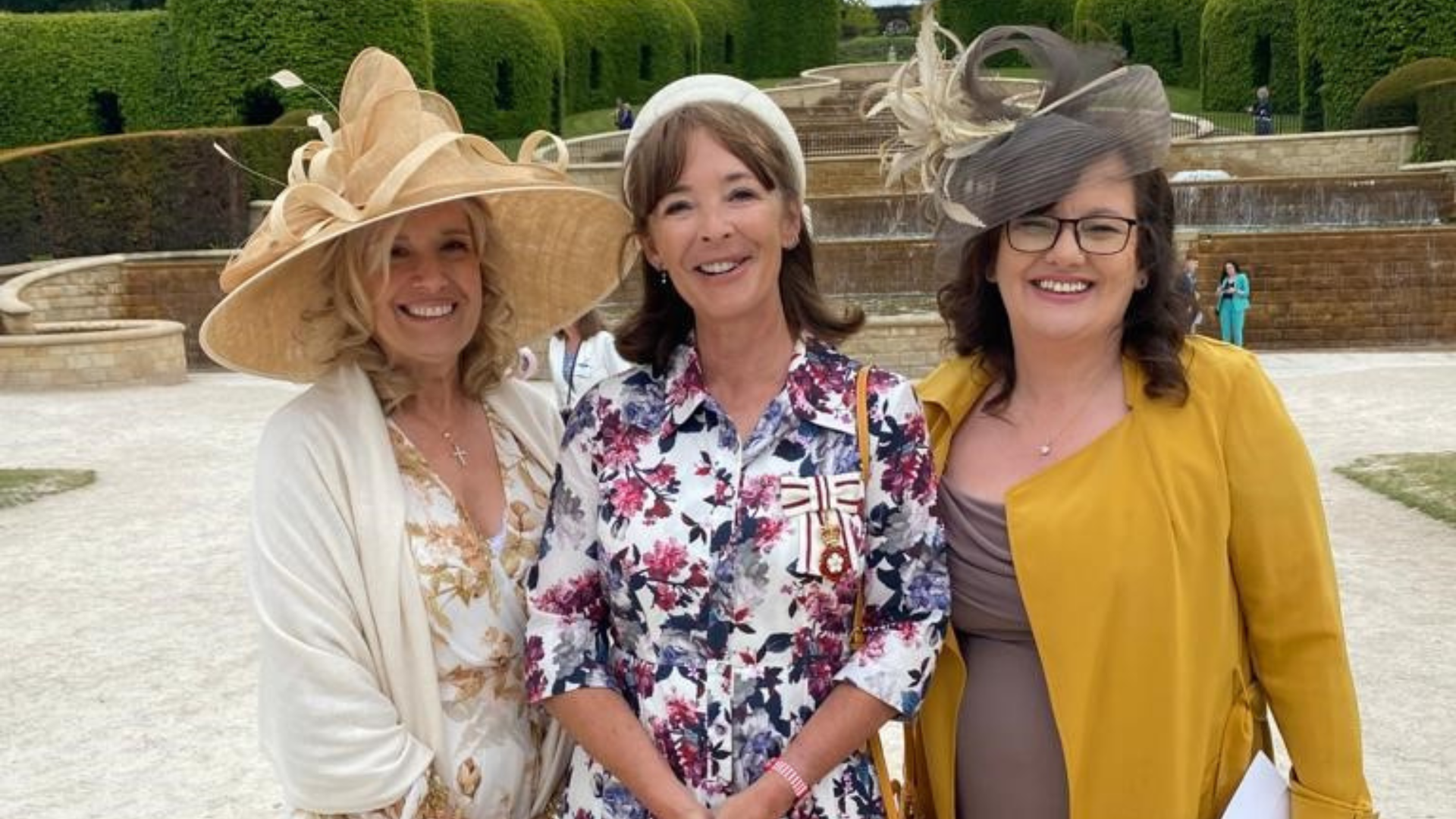 Three ladies dressed in formal dresses and hats smiling at camera