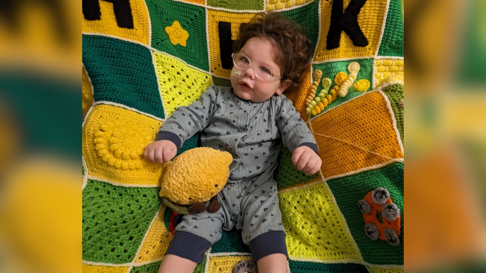 A toddler wearing glasses and laying on a yellow and green knitted blanket