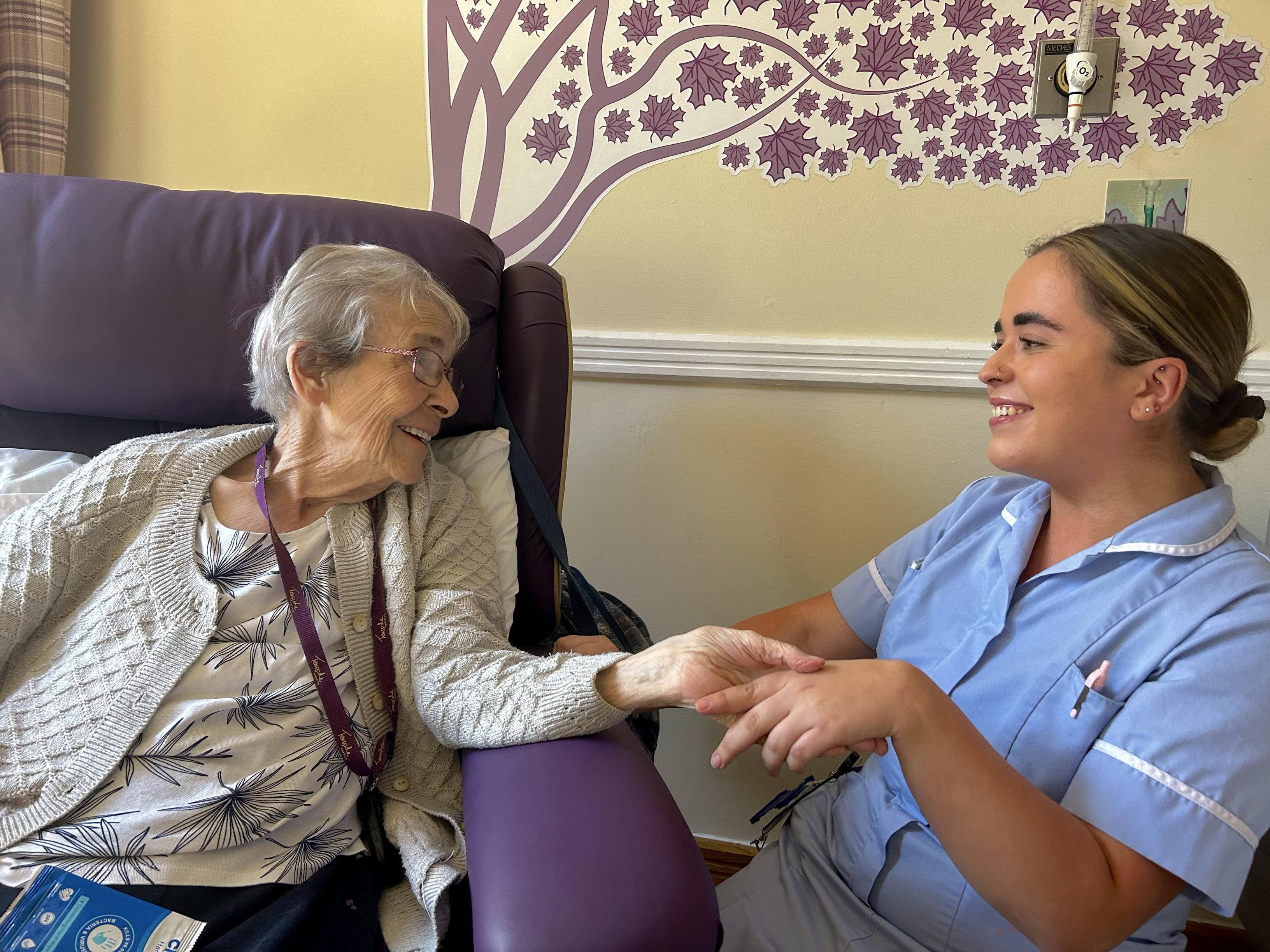 An elderly woman sitting on a winged back chair and holding hands with a nurse