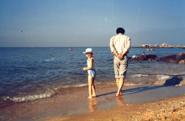 an adult male walks away from the camera on a beach, near the lapping waves. A child next to him, turns to look at the camera