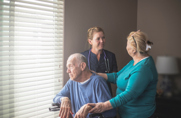 A man in a wheelchair looks out the window with carer and a nurse behind