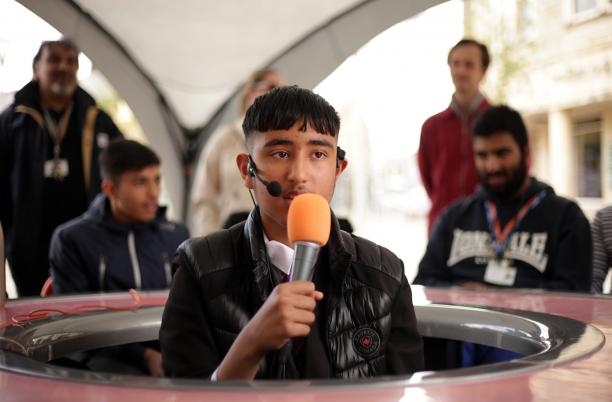 A young man holds a microphone and sits in the middle of a round table, looking outwards and those sitting around the table.