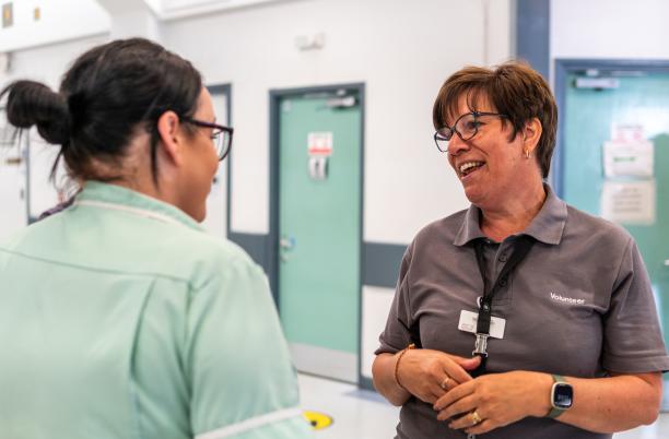 Two women talking to each other in a clinical setting. On the left, one woman is wearing pale green scrubs. On the right, a woman is wearing a grey t-shirt which says 'volunteer'.