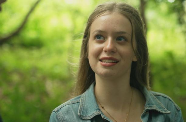 A teenager with long, brunette hair looks up smiling. She is outdoors among the trees of a forest.