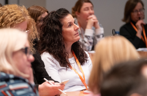 A woman with long dark hair sitting in a crowded room at a conference event.