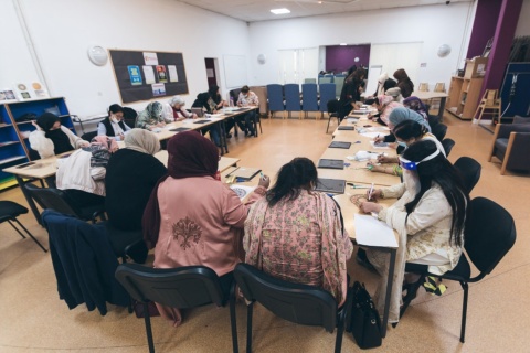 a group of women of sit around a rectangular configuratin of tables, mostly with their backs to the camera. The women appear to be doing a craft activity, and may be drawing.
