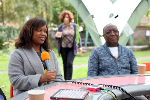 A woman holds a microphone whilst sitting at a round table that is located outside 