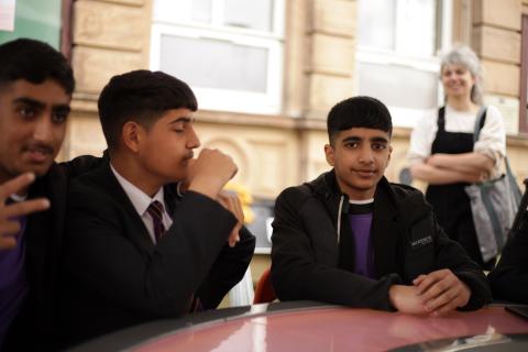 Three young men sit around a round table which is in an outside environment 