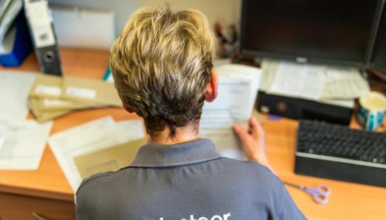 A woman with short blonde hair sitting at a desk with her back to the camera. Her grey t-shirt says 'volunteer' on the back.