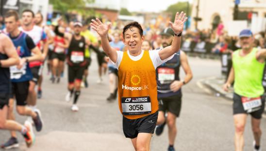 A man running the marathon in an orange branded Hospice UK vest waving to the camera as he runs past.